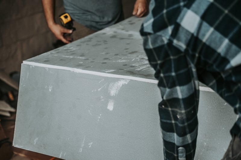 Two workers lift aDensShield Tile Backer board together, one holding it from the foreground and another gripping a drill behind it.