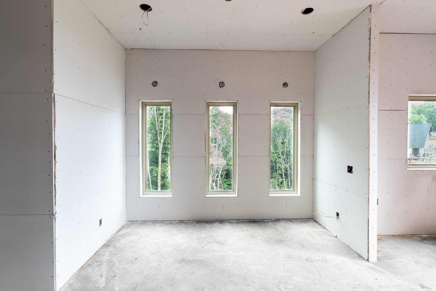 Construction interior showing newly installed white ToughRock® gypsum wallboard panels covering the walls and ceiling, three tall narrow windows in a row on the far wall overlooking green trees, exposed electrical boxes, and an unfinished concrete floor.