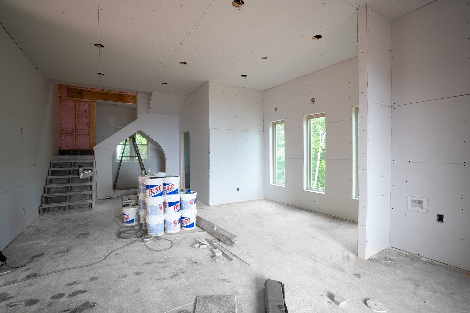 Stack of gypsum wallboard panels with printed edges standing vertically on a concrete floor next to exposed wooden wall studs at a job site.