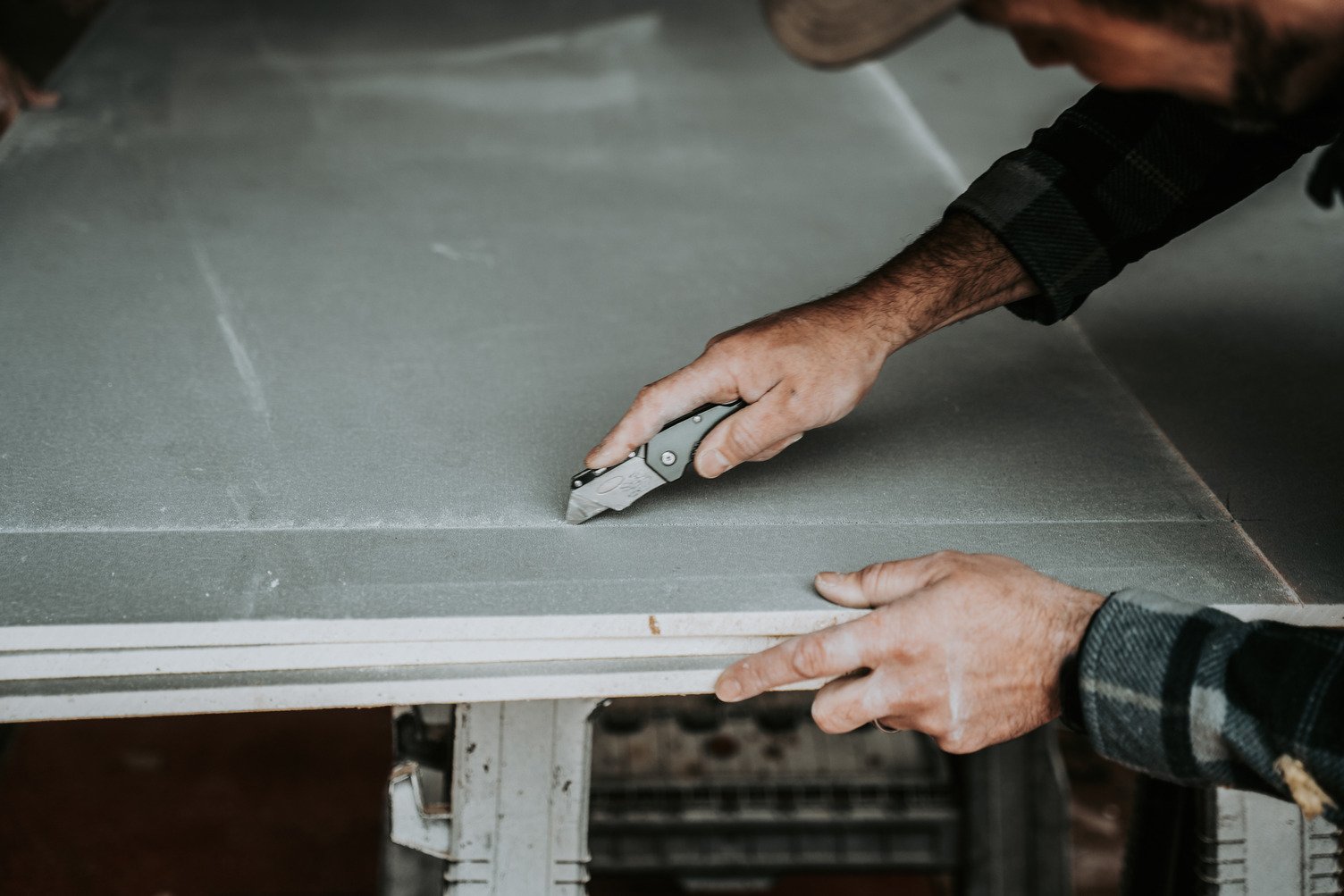 Close-up of a worker’s hands using a utility knife to score and cut a grey DensShield® tile backer panel on a workbench, with part of their arm and plaid shirt visible.