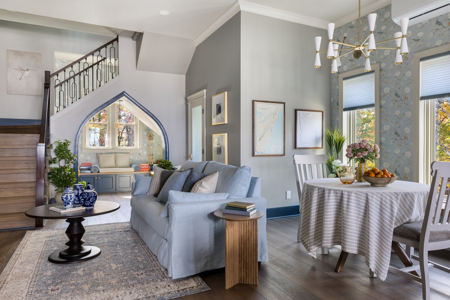 nterior of a stylish open-plan living and dining area featuring a light blue upholstered sofa, a round wood dining table with chairs, floral wallpapered accent wall, and tall windows letting in natural light.