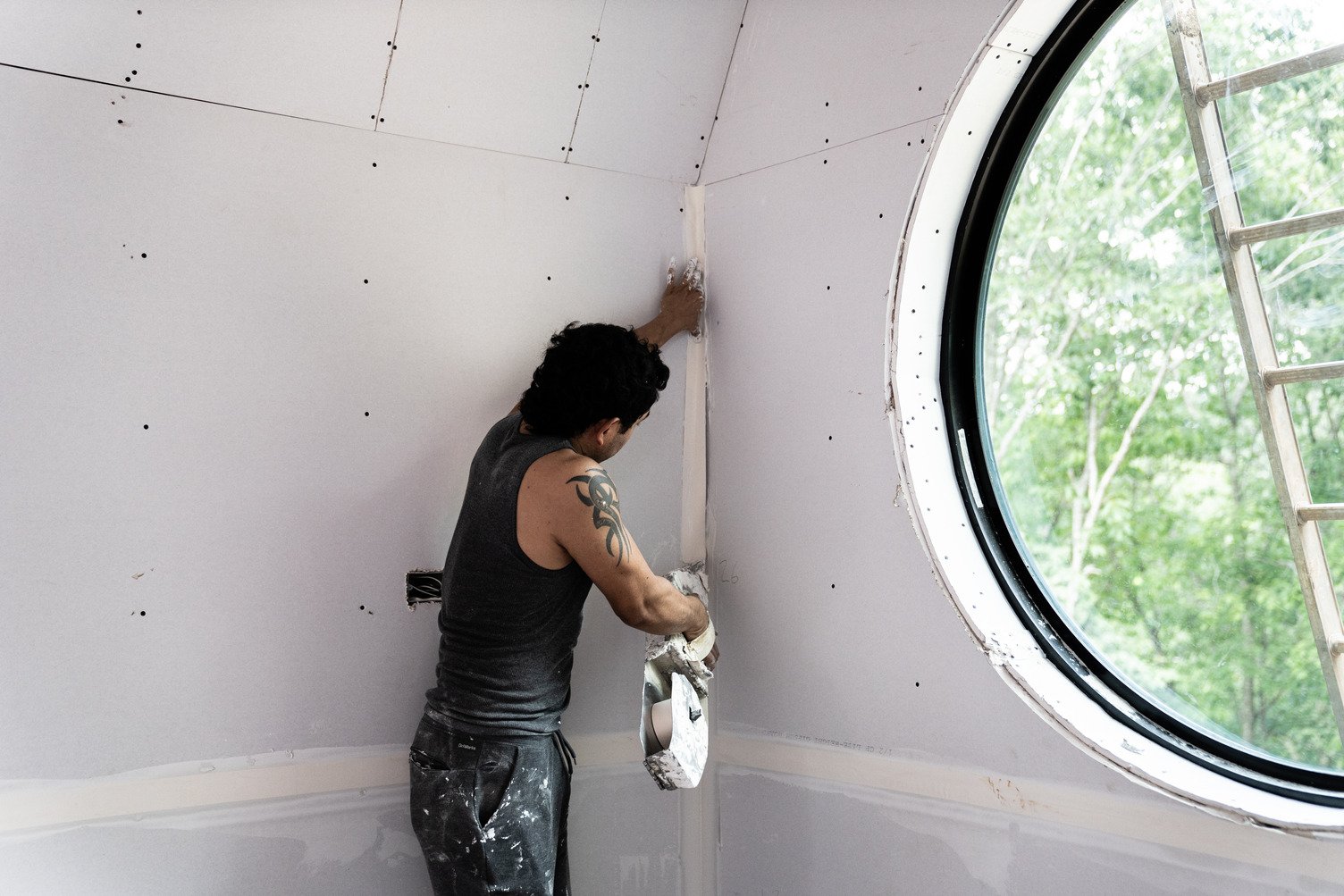Worker on stilts securing gypsum wallboard panels to a high sloped ceiling in an unfinished attic, with exposed wood framing and insulation around them.