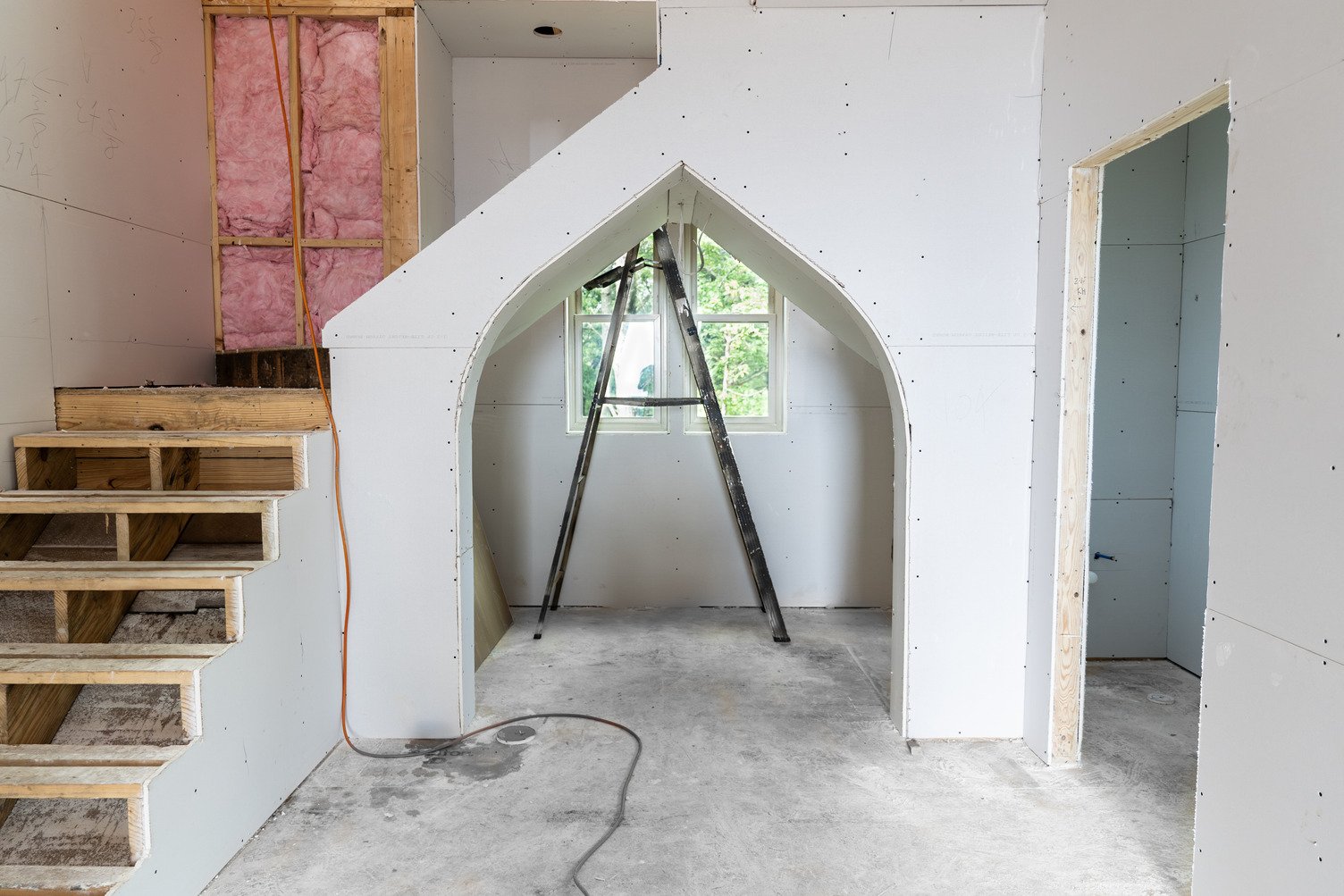 Construction interior with white gypsum wallboard (ToughRock®) installed on framed walls, an arched opening leading to another area with a window and ladder visible, exposed insulation and wood framing on the left, and unfinished wooden stairs.