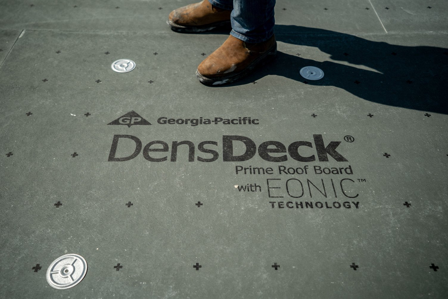 A close-up view of a worker’s boots standing on a rooftop surface printed with ‘Georgia-Pacific DensDeck Prime Roof Board with EONIC Technology’ text and fastening points.