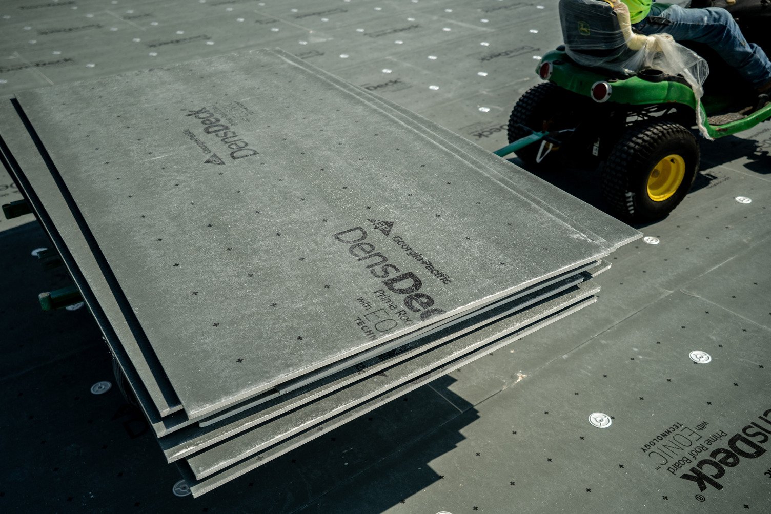 A stack of gray DensDeck Prime roof boards with visible branding placed on a rooftop work area, with a ride-on lawn mower and construction surface in the background.