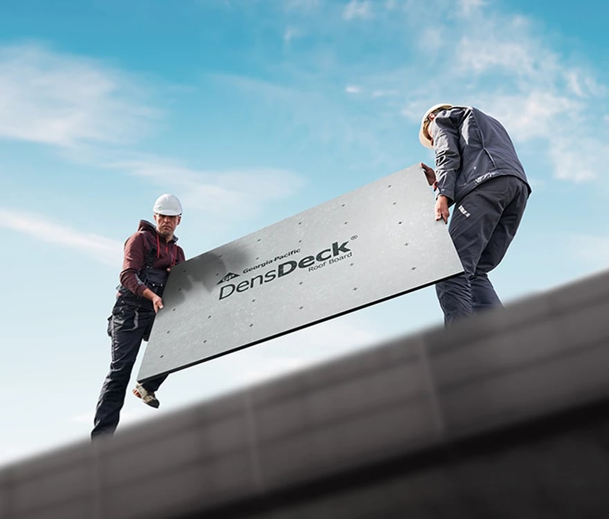 From a lower perspective, we see two roofers above, one wearing gray overalls, a burgundy hoodie and white hard hat, the other wearing gray pants, a dusty blue jacket and a white hard hat, both holding either side of a light gray DensDeck® Roof Board on top of a roof. There is a blue sky and white clouds in the background.