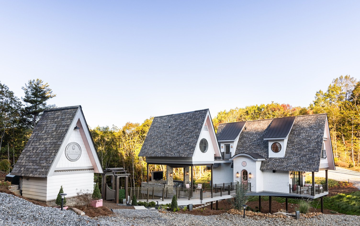 Photo of three white and gray mountain treehouses with steep gabled roofs and round windows, elevated on supports above gravel and surrounded by forest.