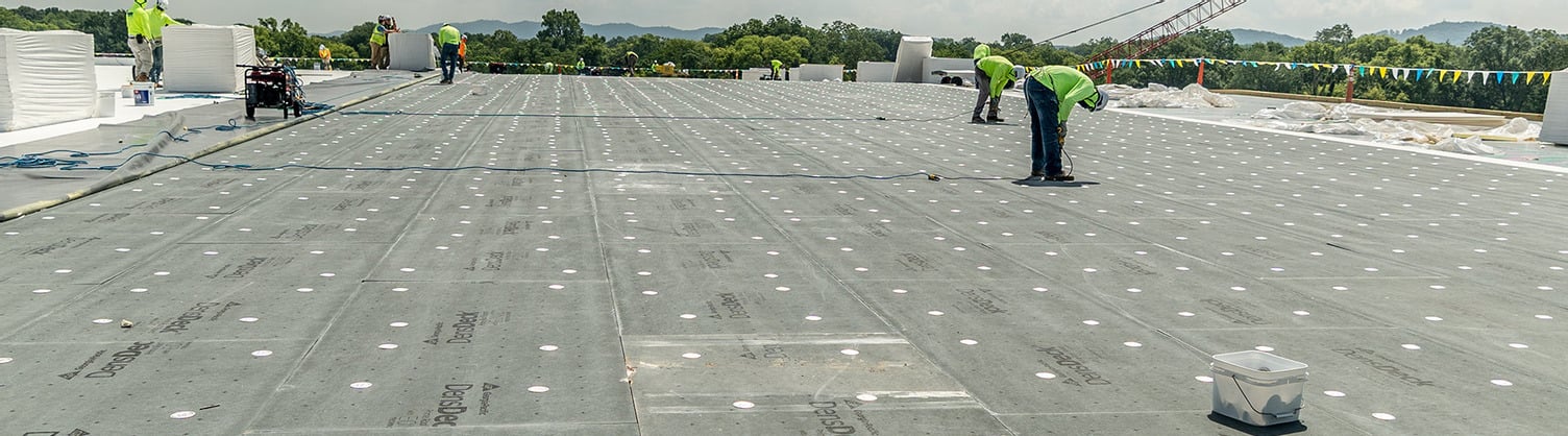 Wide panoramic view of workers installing DensDeck roof boards on a large flat commercial roof at the Coca-Cola warehouse construction site in Alabama, with construction materials and safety gear visible under a partly cloudy sky.