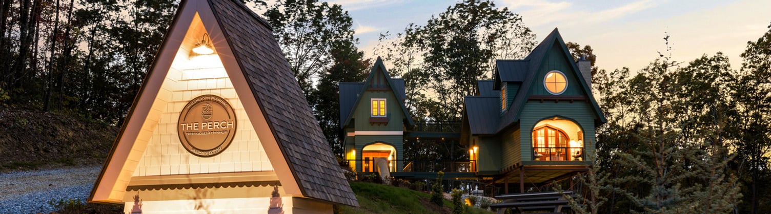 Wide exterior shot of modern elevated treehouse structures with steep gabled roofs, raised on stilts above a gravel driveway and surrounded by wooded hills.
