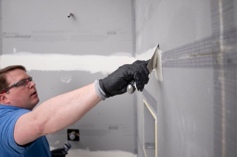 Man in a blue shirt and safety glasses spreading joint compound along a taped seam on a gray DensShield® tile backer board wall in a bathroom installation.