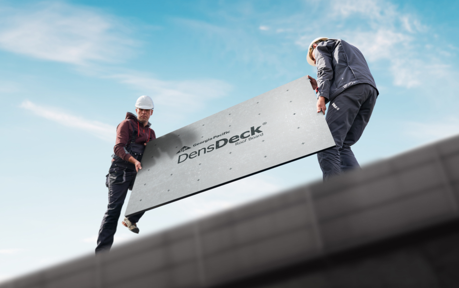 From a lower perspective, we see two roofers above, one wearing gray overalls, a burgundy hoodie and white hard hat, the other wearing gray pants, a dusty blue jacket and a white hard hat, both holding either side of a light gray DensDeck® Roof Board on top of a roof. There is a blue sky and white clouds in the background.