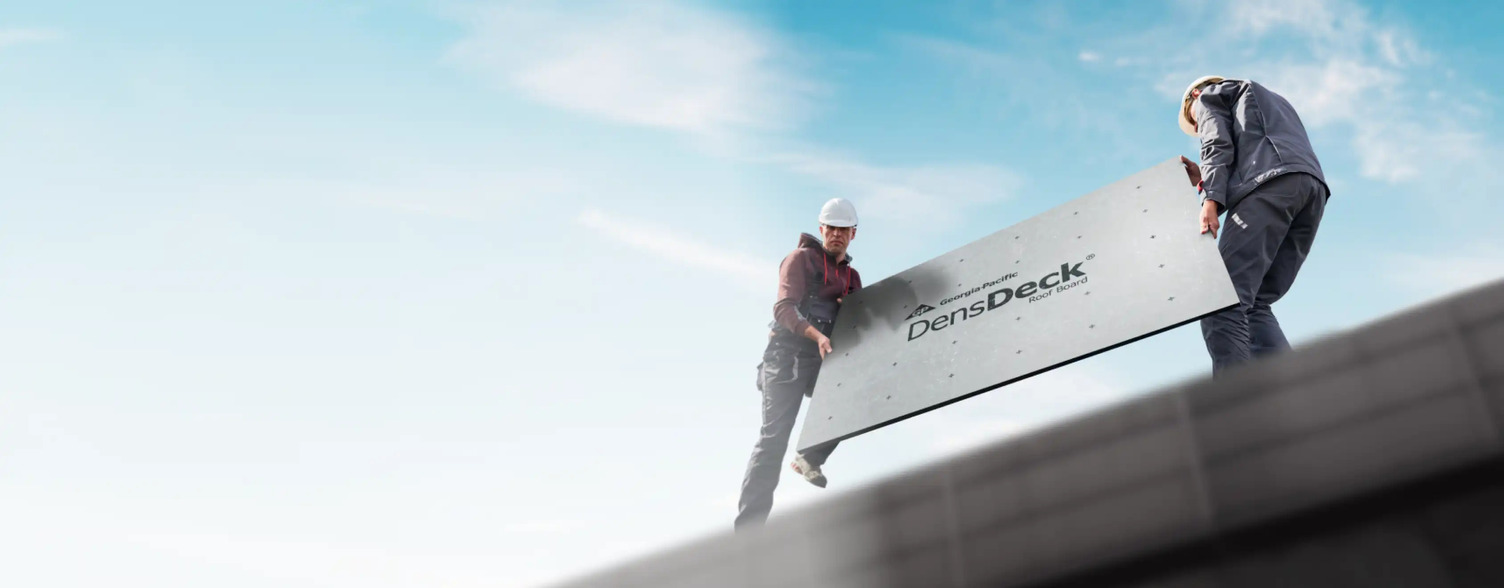 From a lower perspective, we see two roofers above, one wearing gray overalls, a burgundy hoodie and white hard hat, the other wearing gray pants, a dusty blue jacket and a white hard hat, both holding either side of a light gray DensDeck® Roof Board on top of a roof. There is a blue sky and white clouds in the background.